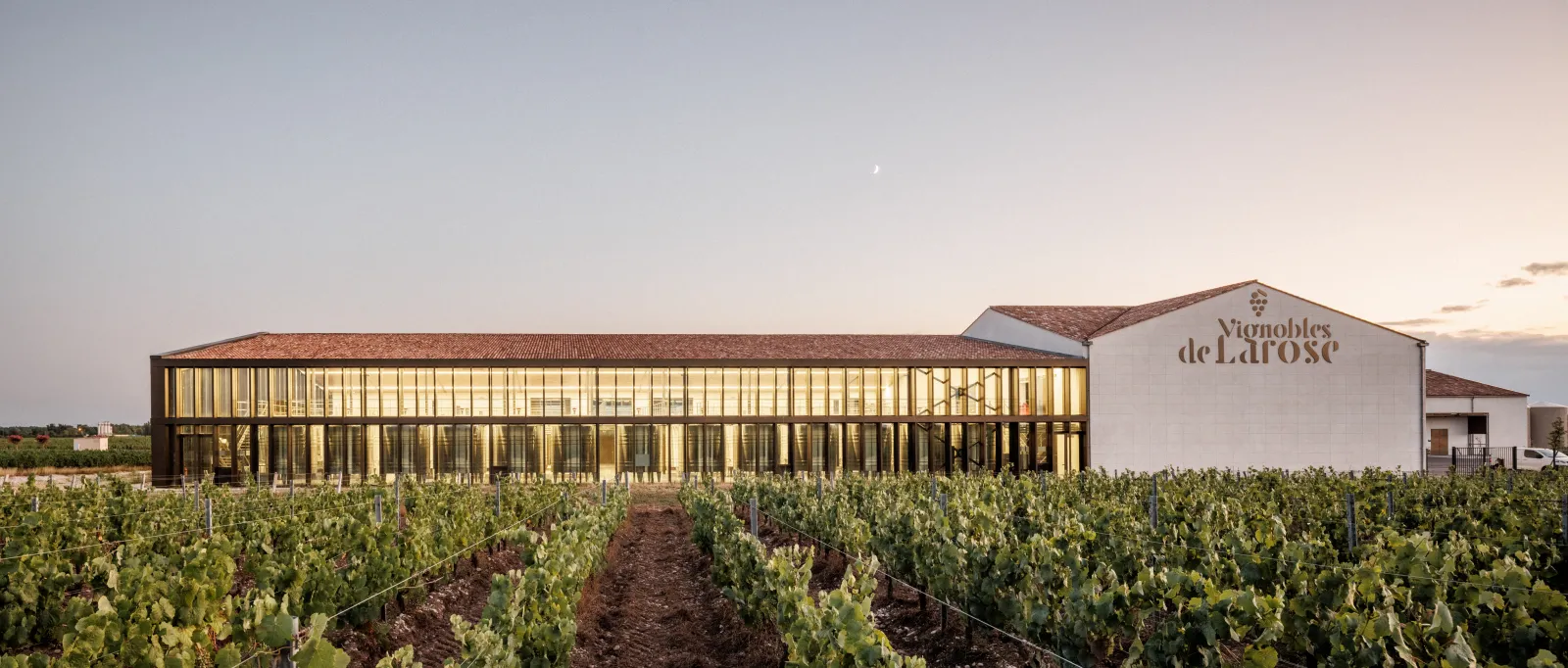 Modern architecture of Château Larose-Trintaudon in Bordeaux with golden glass and metal facades, surrounded by vineyard rows at sunset.