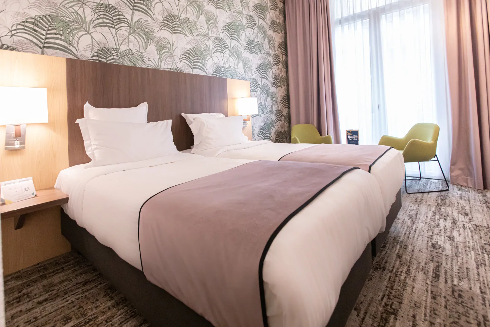 Modern hotel room with double bed featuring white sheets and pink bedspread, wooden headboard, bedside lamps, floral patterned wallpaper, olive green armchair, pink curtains and window view at Quality Hôtel Bordeaux Centre.