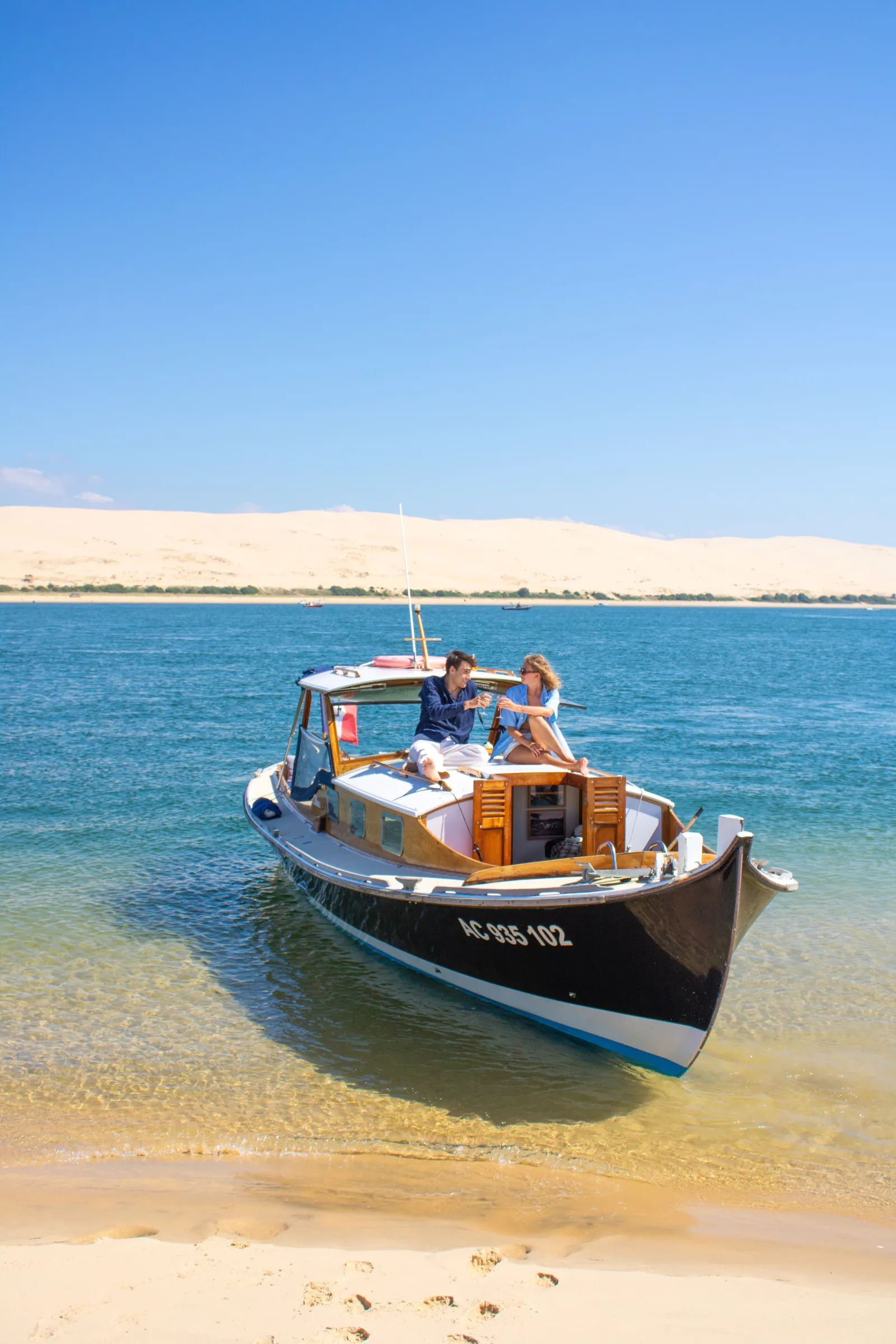 Two people aboard a traditional wooden fishing boat anchored in shallow turquoise water with a white sand dune in the background.