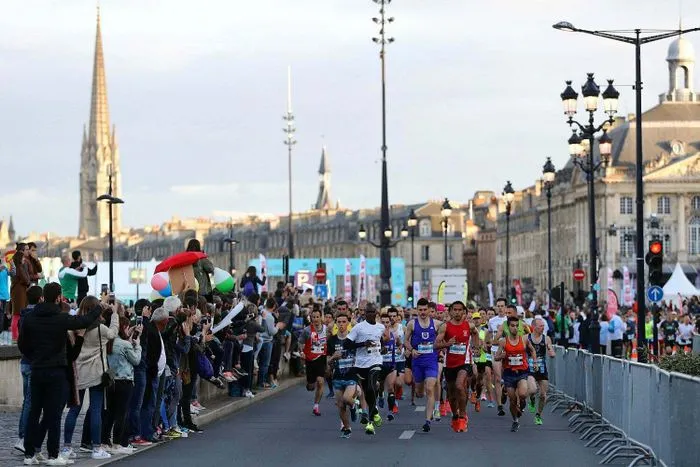 Runners participating in the Bordeaux half-marathon, running on a closed road with spectators lining the course and historic Bordeaux architecture in the background.