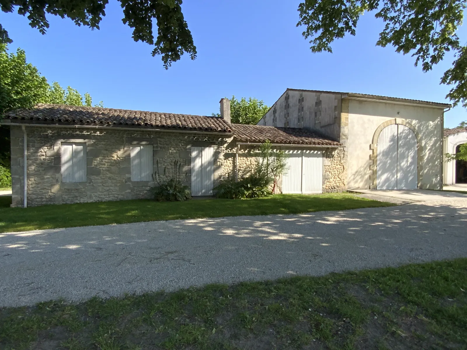 Stone facade of Château d'Osmond in Bordeaux featuring white doors, paved driveway, and surrounding greenery under clear blue sky.