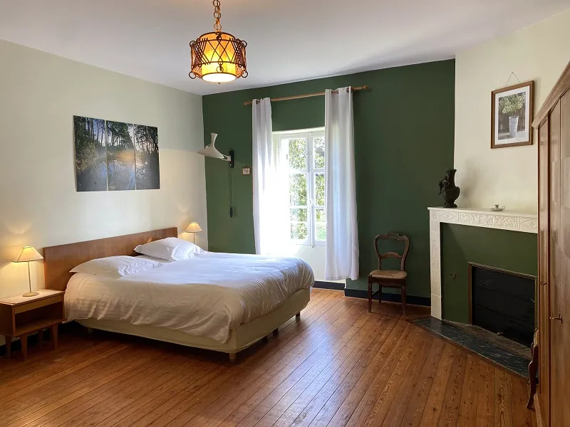 Elegant bedroom featuring a wooden king-size bed, dark green and cream walls, white fireplace, French doors with white curtains overlooking a garden, light hardwood flooring, and vintage gold ceiling fixture.
