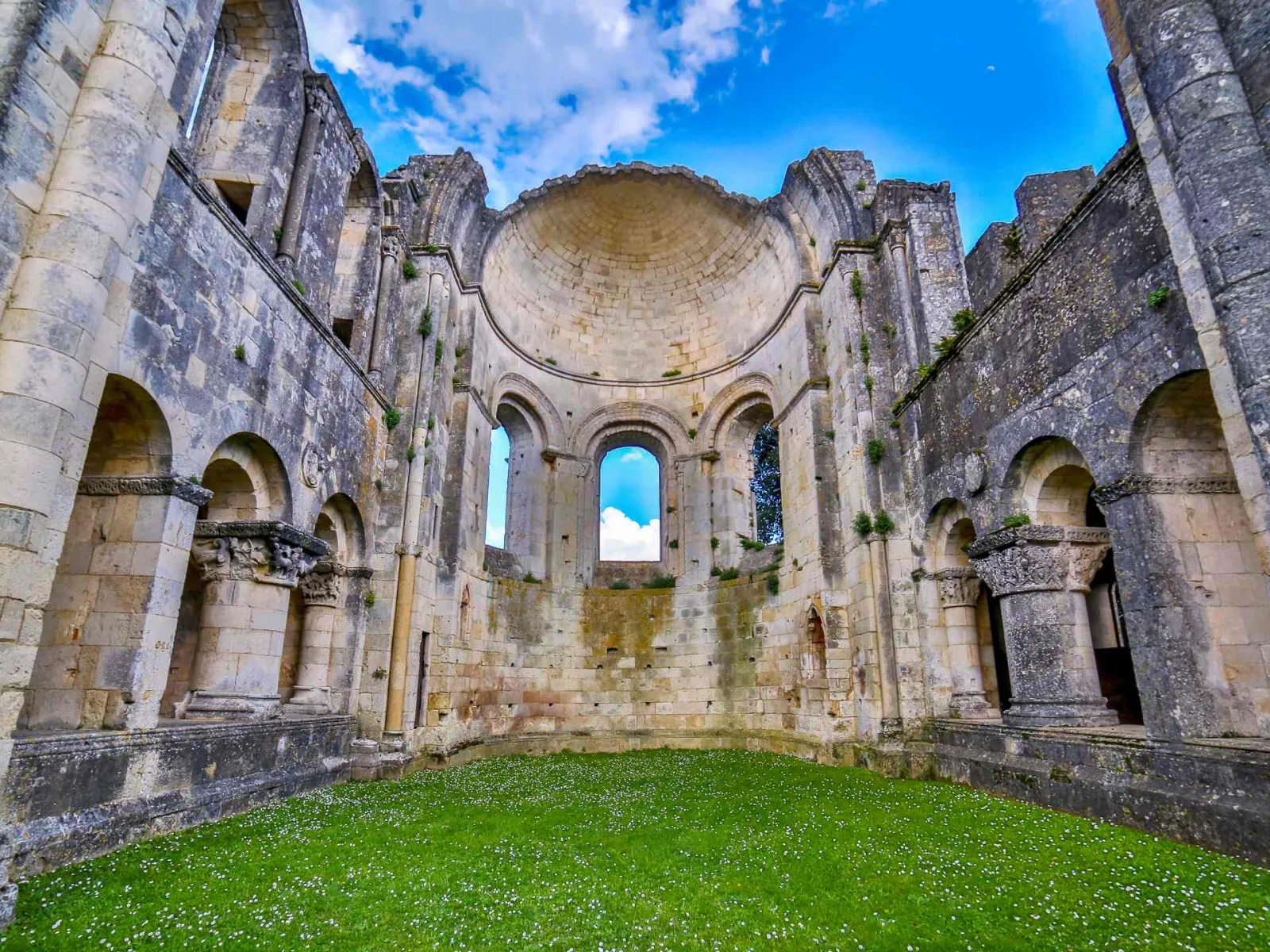 Ruins of La Sauve-Majeure Abbey near Bordeaux, featuring the interior of a Romanesque stone church with barrel vault, columns with capitals, rounded arches and green lawn beneath a clear blue sky.