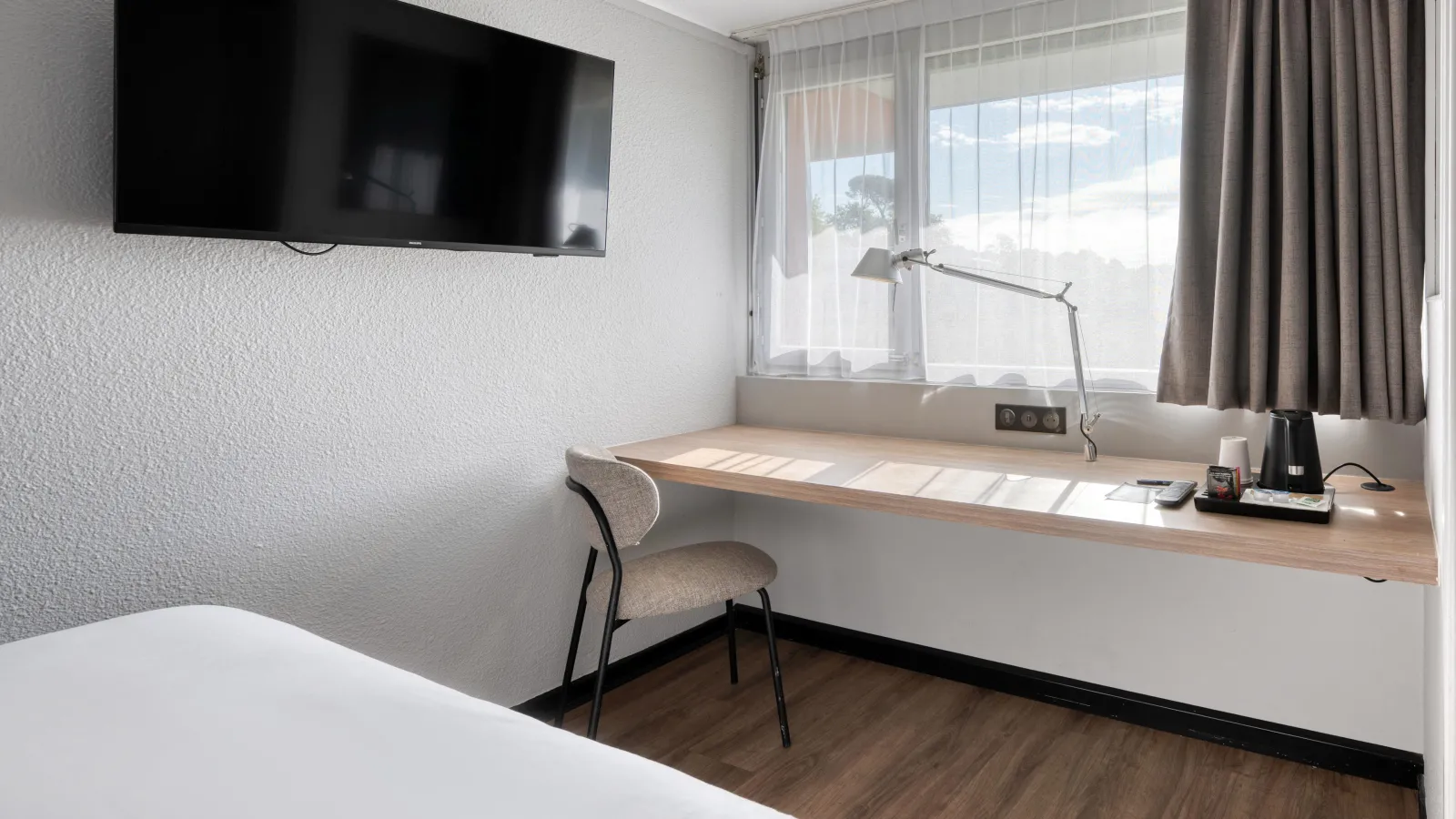 Modern hotel room featuring a wooden desk by a large window, beige chair, wall-mounted flat-screen TV, desk lamp, and sunny outdoor view in Bordeaux