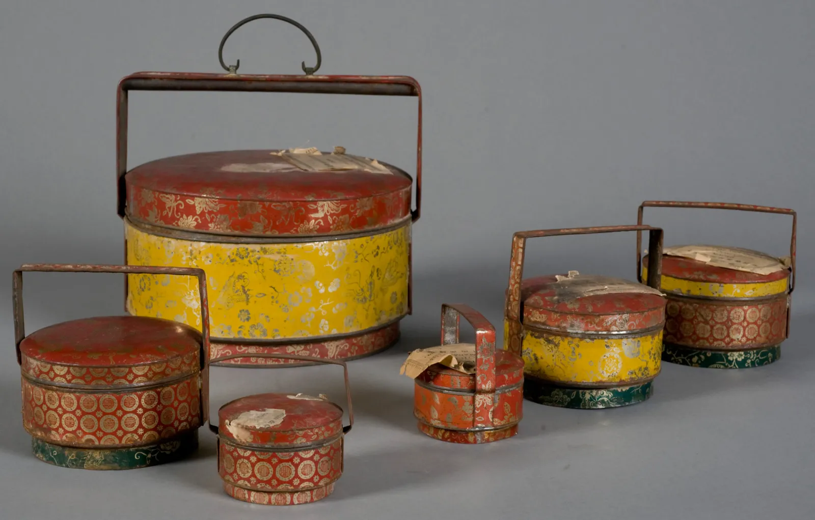 Collection of Chinese tiered lacquerware boxes in red and yellow with gold floral and geometric patterns, equipped with metal handles, displayed at the Bordeaux Museum of Ethnography.