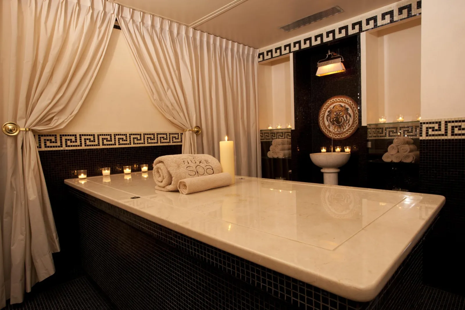 Luxurious bathroom at Guerlain Spa in Bordeaux featuring a white rectangular bathtub, black and white geometric tile work, beige curtains, Mediterranean-inspired décor, and lit candles creating a serene spa atmosphere.