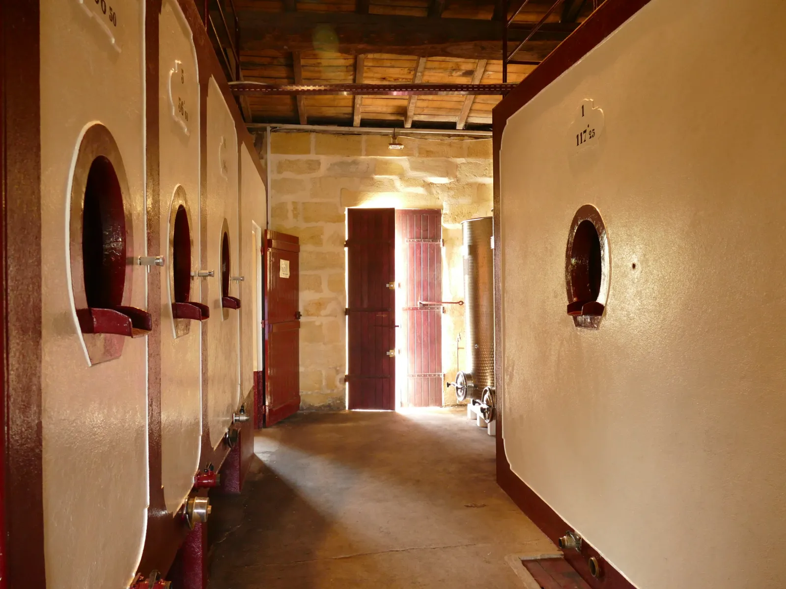 Wine storage corridor at Château Laniote in Bordeaux featuring wooden barrels lined along cream and burgundy walls with red doors and natural light
