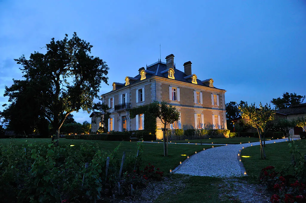Château Haut-Bailly lit up at dusk, an elegant 19th-century Bordeaux mansion with ochre facades and slate roofs, surrounded by vineyard grounds, centuries-old trees, and an illuminated gravel pathway leading to the entrance.