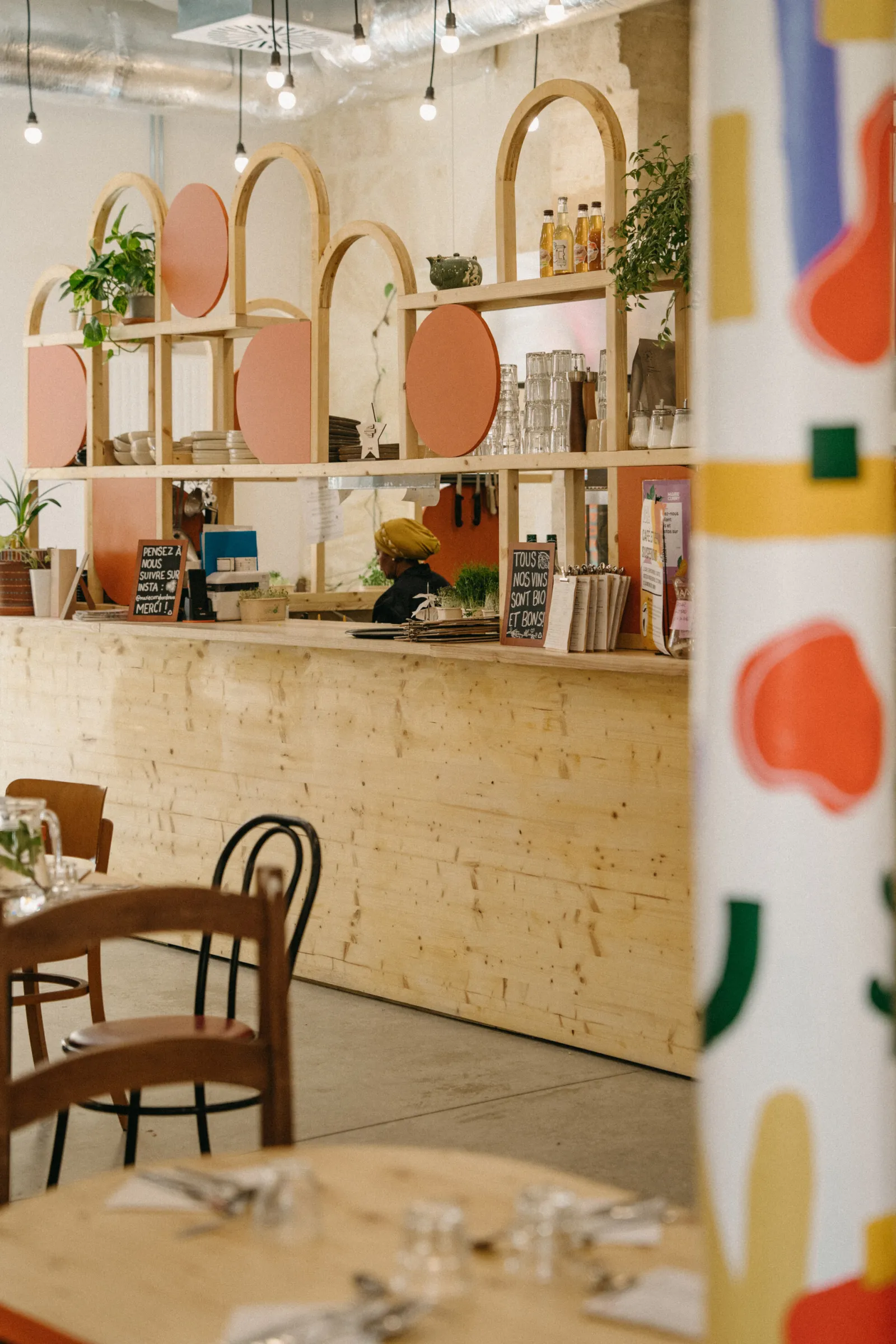 Interior of Marie Curry restaurant in Bordeaux featuring a light wood counter, decorative shelves with pink mirrors and gold arches, potted plants, wooden chairs, and colorful wall accents.