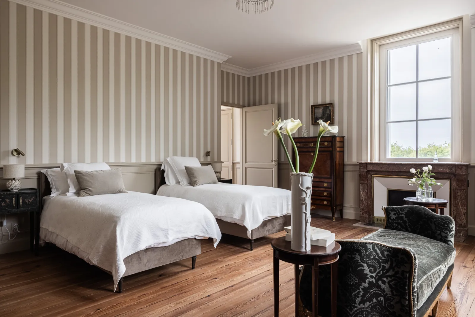 Elegant guest bedroom at Château Sénéjac in Bordeaux featuring two twin beds with white linens, beige striped wallpaper, marble fireplace, antique furniture and large windows.