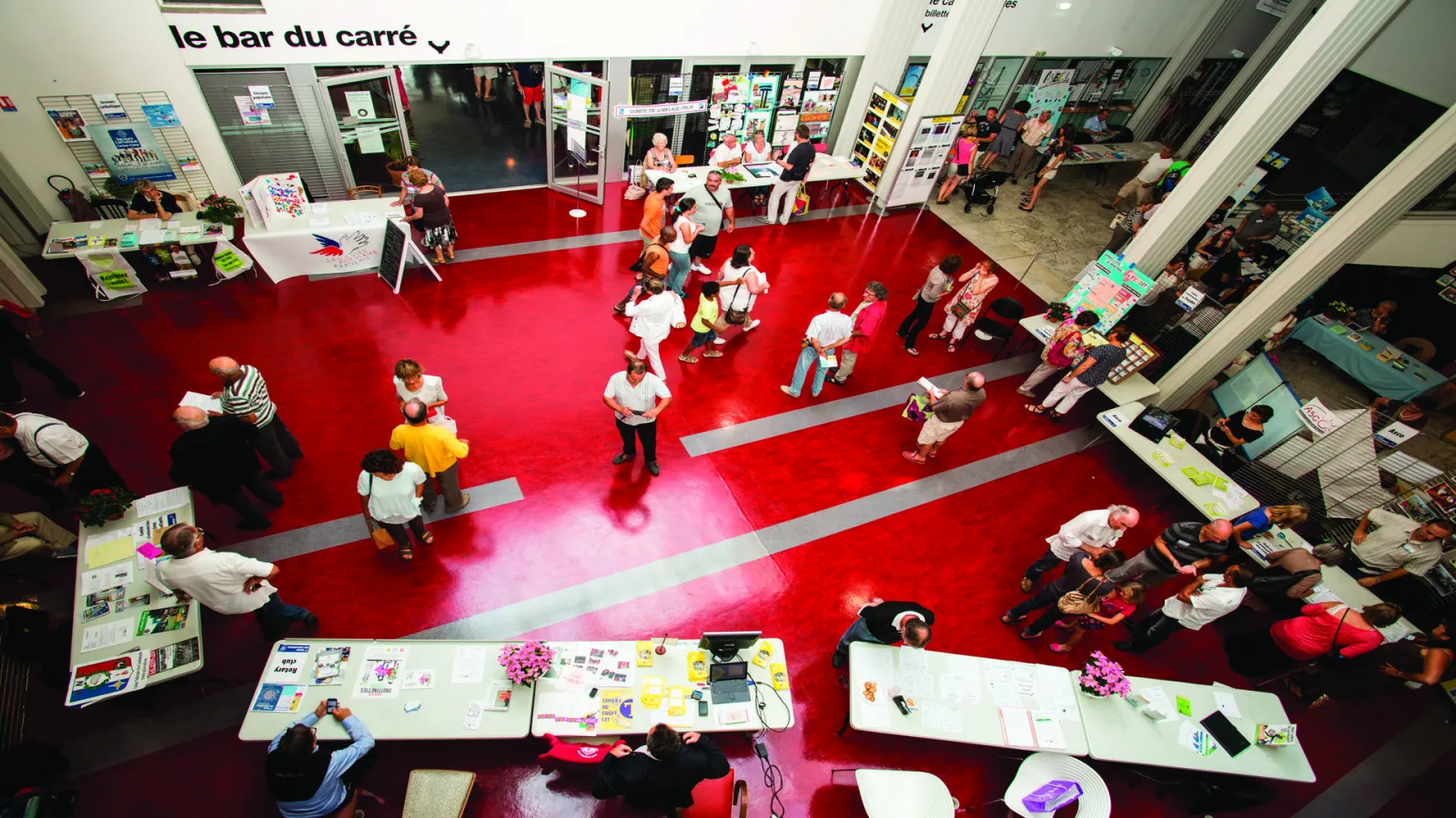 Overhead view of le bar du carré in Saint-Médard-en-Jalles, Bordeaux, featuring a vibrant red floor, white display tables with creative projects, and visitors exploring the cultural venue.