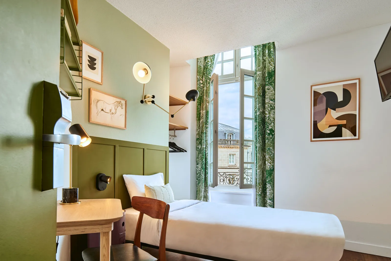 Modern hotel room featuring olive green walls, white bed with upholstered headboard, light wood furniture, contemporary lighting, abstract artworks, French doors opening to a balcony overlooking Bordeaux cityscape with green curtains.