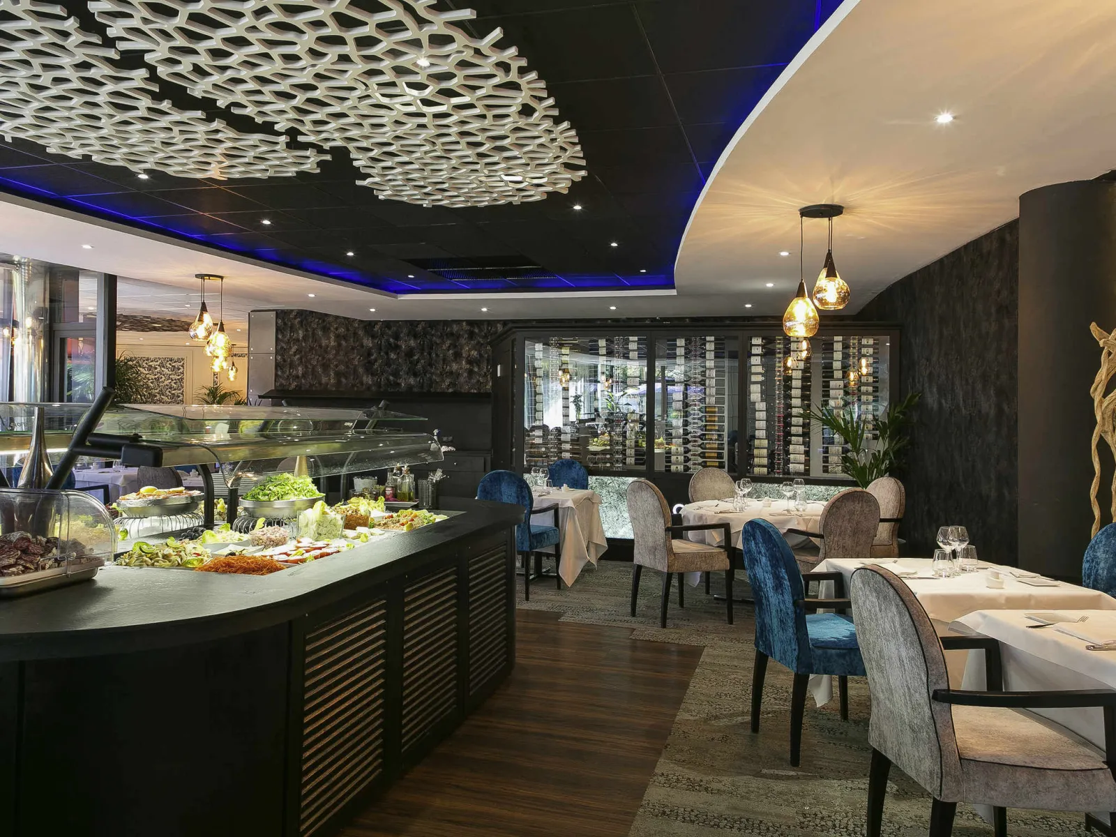 The Spirit's fine dining restaurant in Bordeaux featuring an open kitchen counter, elegant tables, modern black and gold décor, geometric white ceiling, and glass wine cellar.