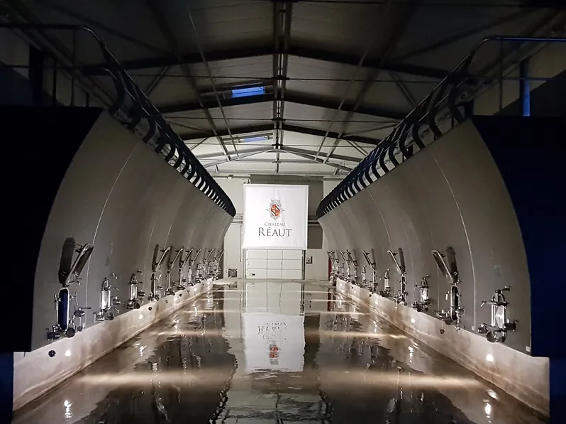 Interior of a symmetrical fermentation tank at Château Réaut in Bordeaux, lined with tasting taps on both sides and the estate's sign at the far end