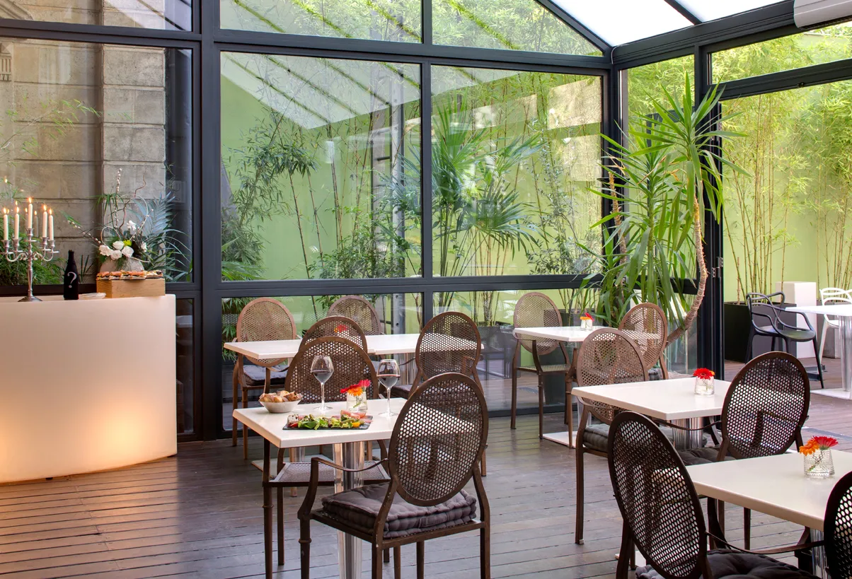 Elegant covered terrace of Bordeaux's Boutique Hotel Bar featuring white tables, black rattan chairs, glass doors overlooking a lush green garden, and minimalist modern décor.