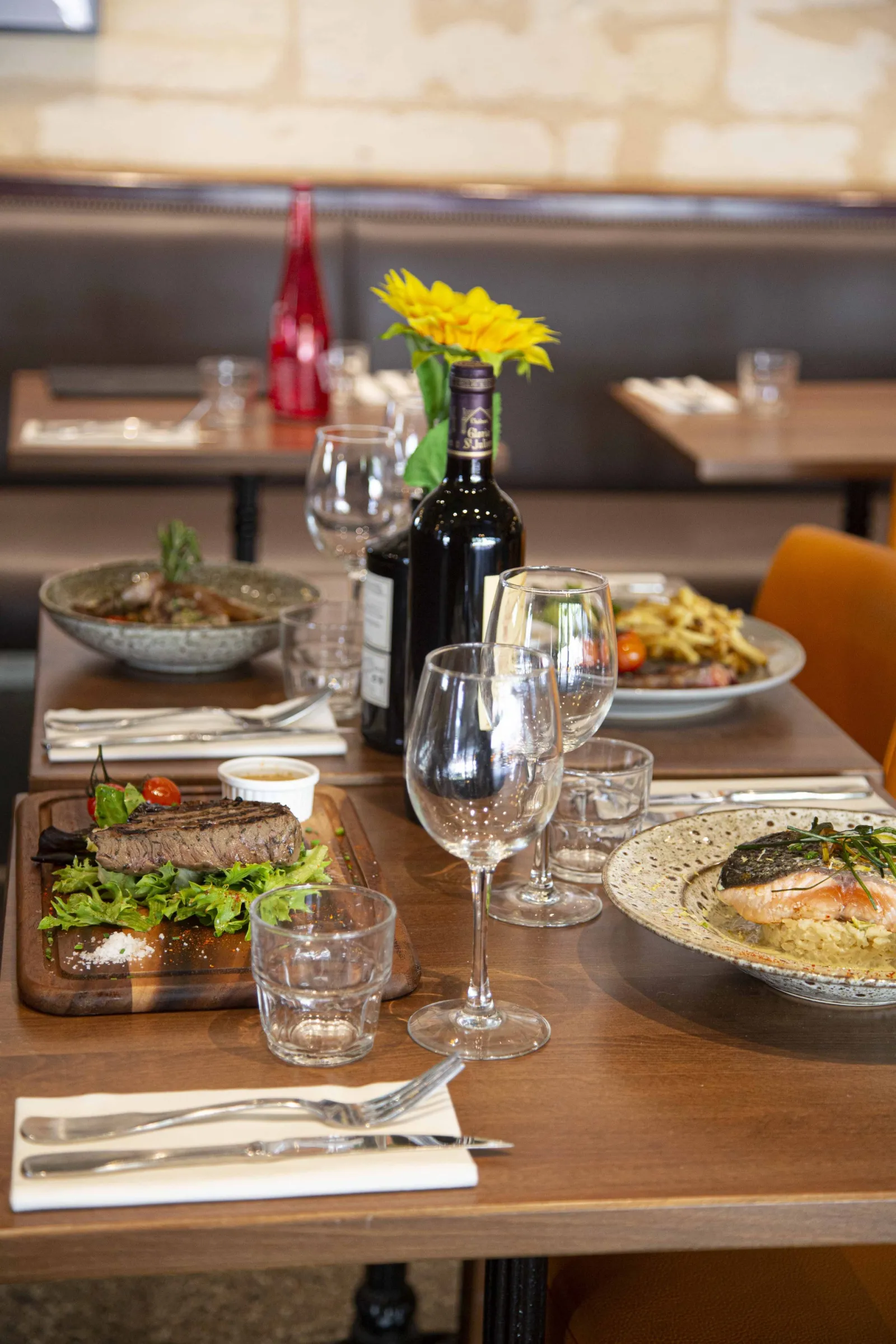 Elegant restaurant table set with gourmet dishes, a glass of red wine, water glasses, cutlery and a yellow flower centerpiece at Cajou Caffé restaurant in Bordeaux.