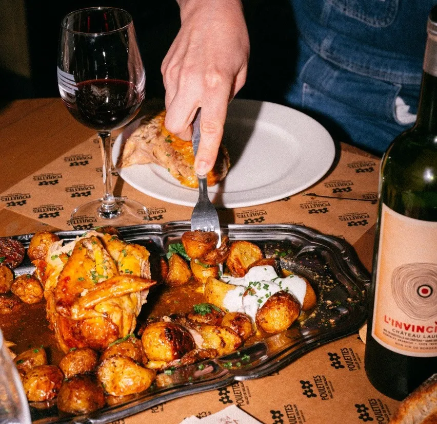A person enjoys roasted chicken with golden potatoes at Rôtisserie Poulettes in Bordeaux, paired with a glass of red wine and a bottle of Pauillac.