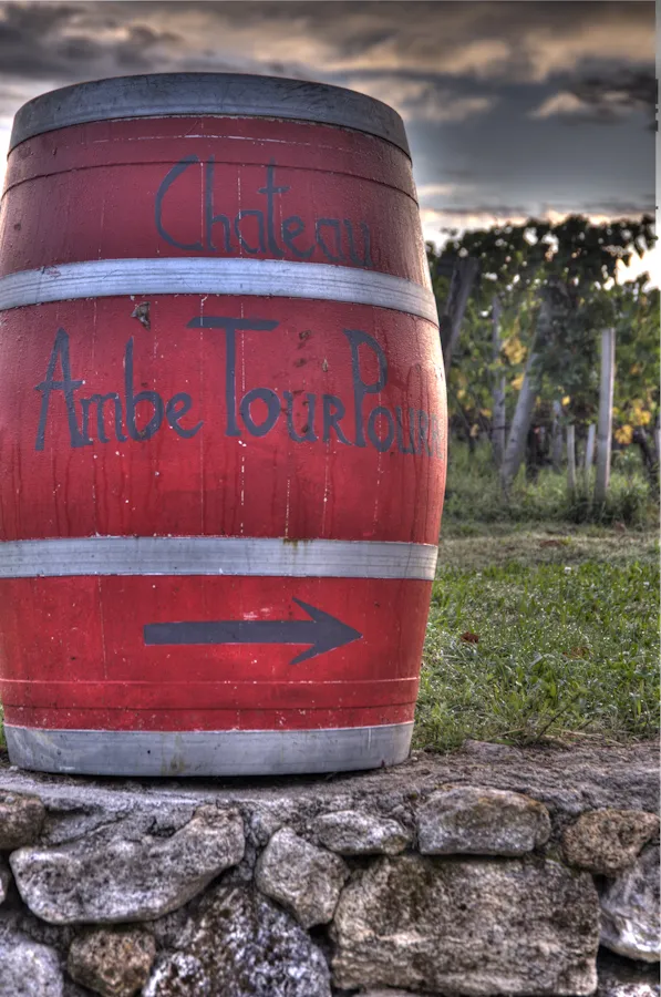 Red painted wine barrel with directional arrows and text markings, placed on a stone wall, with Château Ambe Tour Pourret vineyards visible in the background near Bordeaux
