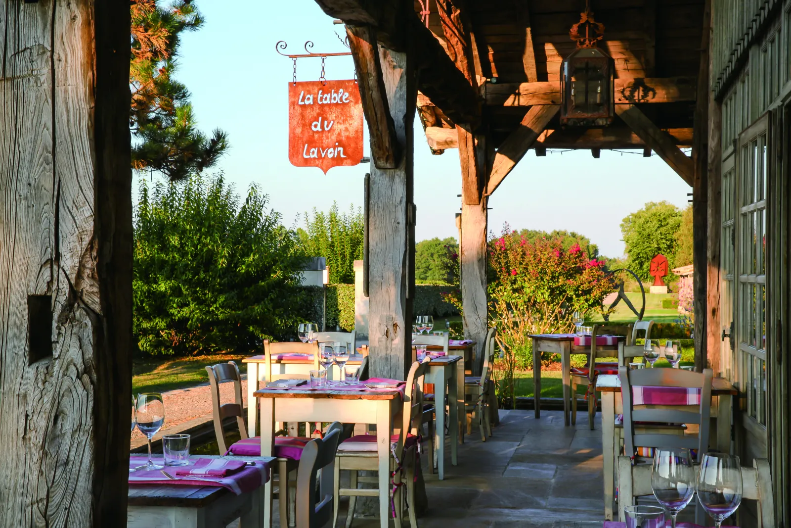 Shaded terrace of La Table du Lavoir in Bordeaux with set tables, purple chairs and red signage, landscaped garden in the background