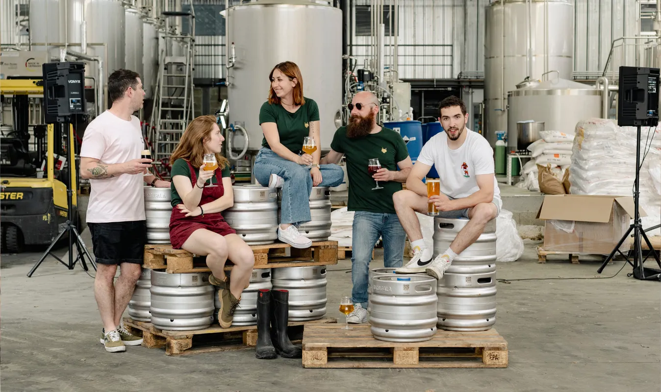 Five people visit Burdigala brewery in Bordeaux, sitting on stacked beer barrels and sampling glasses of beer in the production facility.