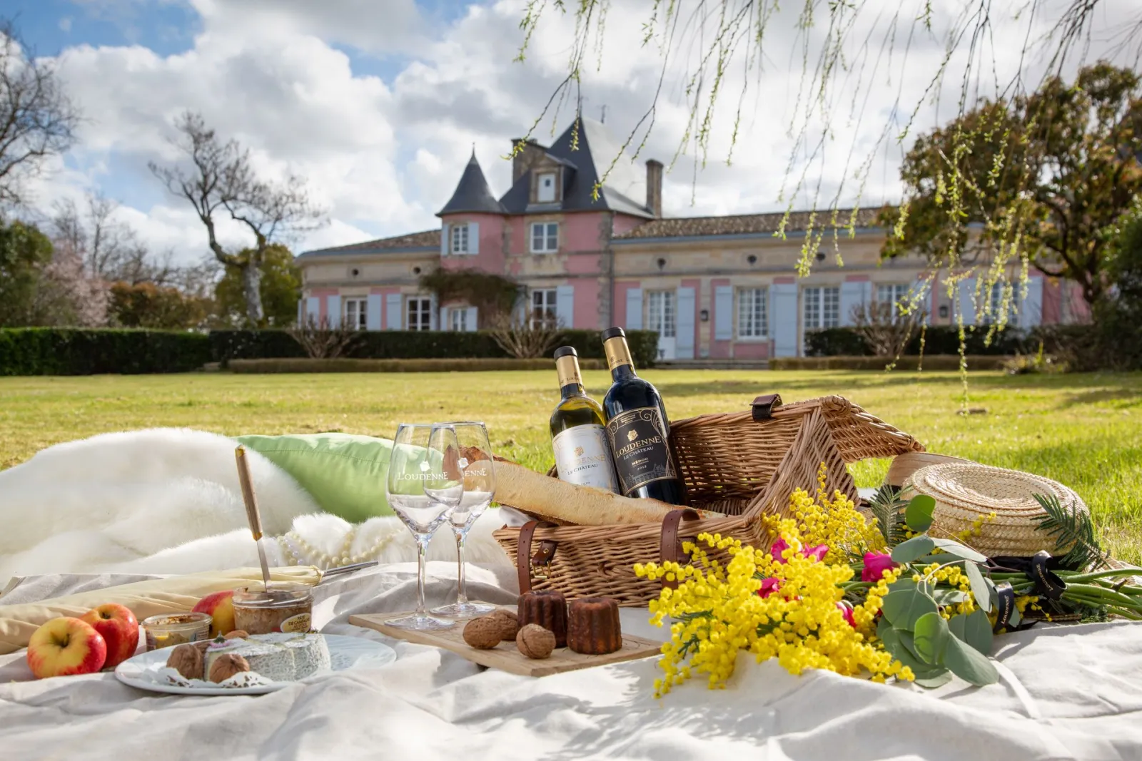 Elegant picnic on a white blanket in the grounds of Château Loudenne in Bordeaux, featuring wine bottles, glasses, fresh fruits, yellow and pink flowers, with the pink château visible in the background.