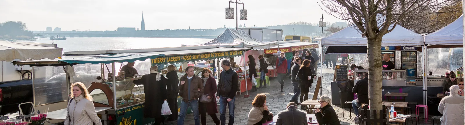 Lively market at Quai des Chartrons in Bordeaux featuring local product stalls, visitors browsing, white market tents, and the Garonne river with the city skyline in the background.