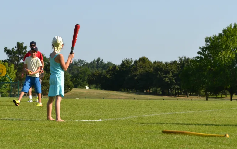 Child holding a red baseball bat on a grass sports field at Domaine du Pinsan in Bordeaux, with adults supervising and other children in the background.