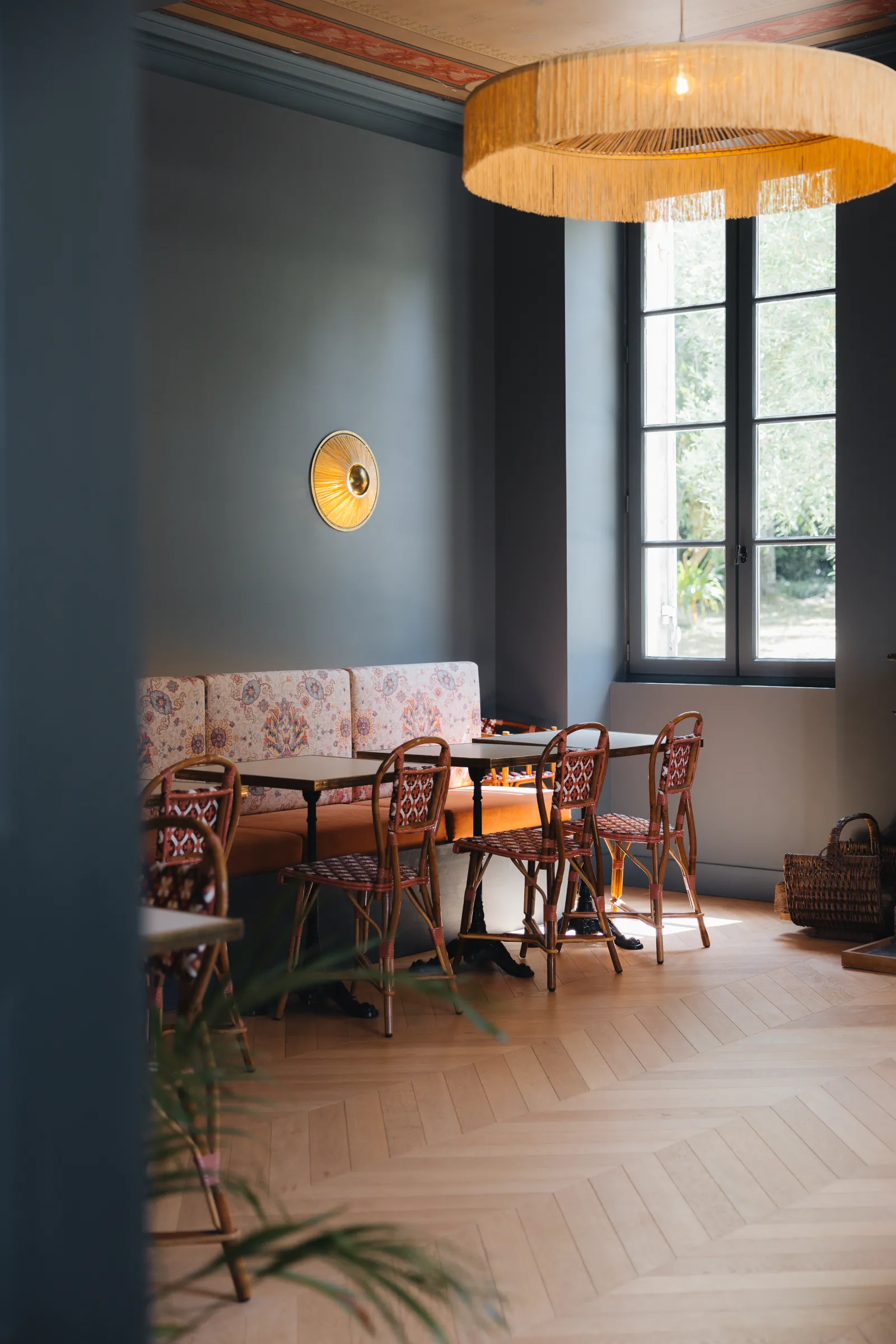 Elegant dining room at Château Réal in Bordeaux featuring a blush pink banquette, rattan chairs, wooden round table, wooden pendant light fixture, blue-gray walls, French window panes, and a brass wall sconce.