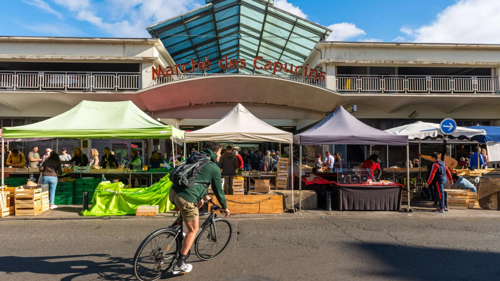 View of the Marché des Capucins in Bordeaux with colorful market stalls, vendors and customers, a cyclist passing by, and the main entrance with its glass canopy in the background.