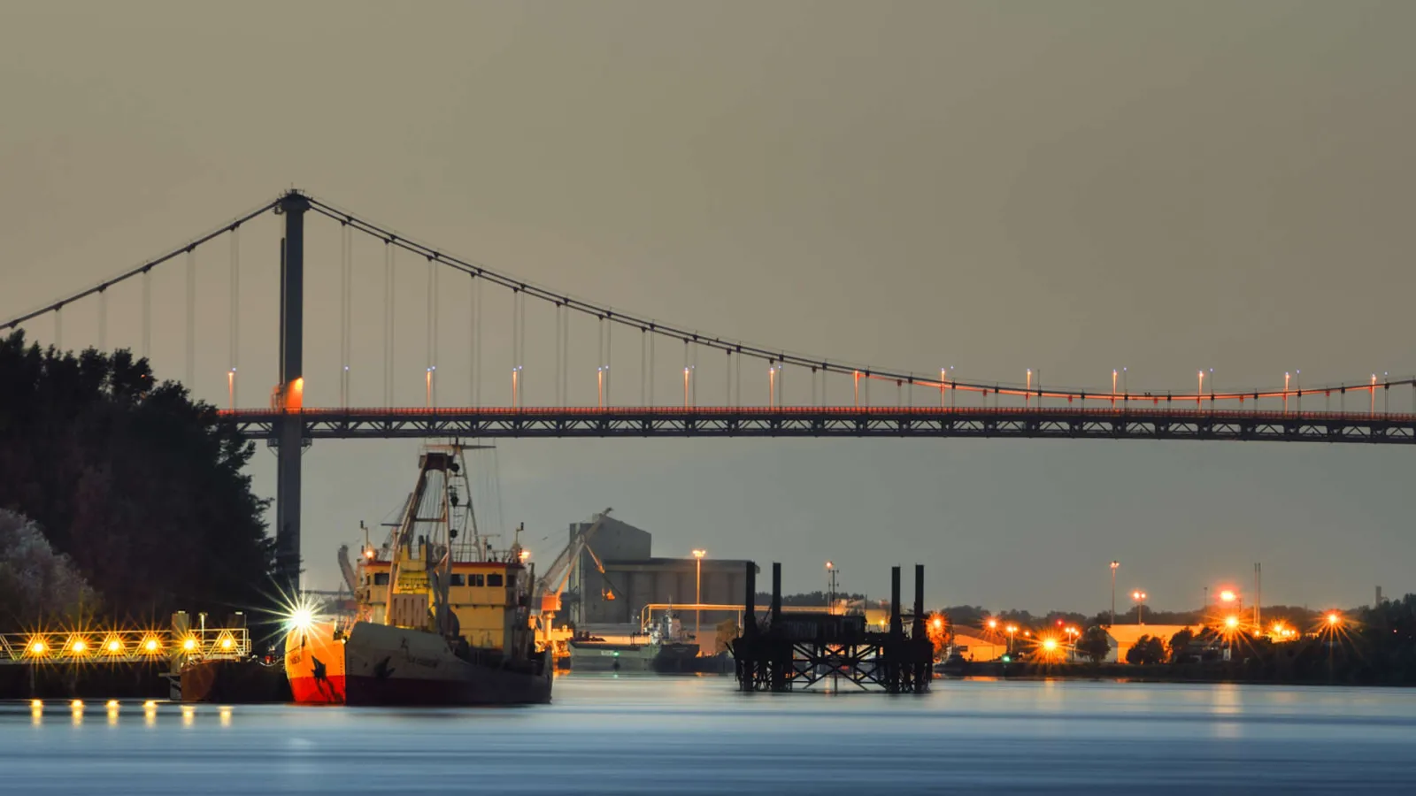 Night view of the Aquitaine Bridge in Bordeaux with an illuminated dredging vessel passing beneath the suspension bridge over the Garonne River, with lit port facilities in the background.