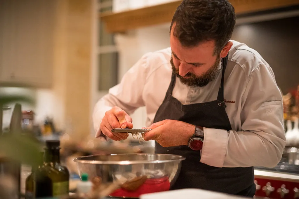 A chef in Bordeaux prepares a dish by pouring a grated ingredient into a stainless steel bowl in his professional kitchen.