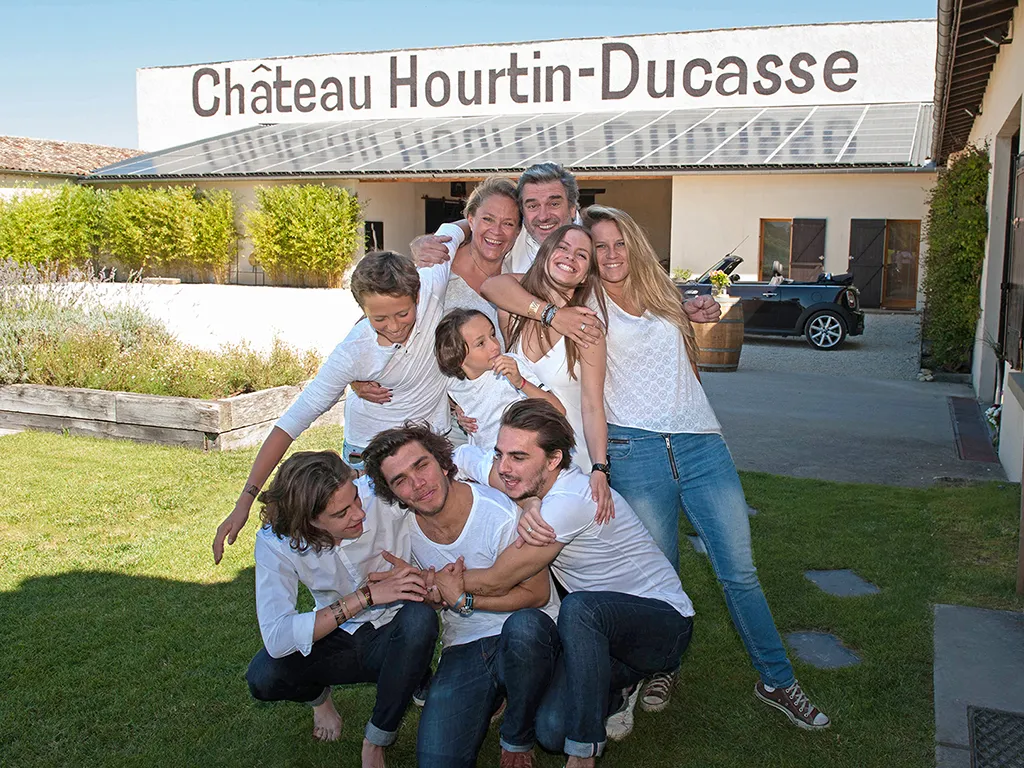 Nine smiling people posing together on the lawn in front of Château Hourtin-Ducasse in Bordeaux, with the main building visible in the background.