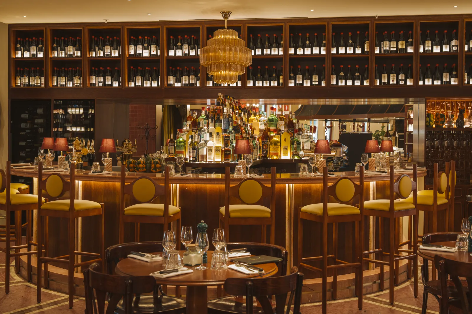 Elegant interior of Brasserie Vinatier in Bordeaux featuring wooden bar counter, yellow chairs, wall-mounted wine cellar, golden chandelier, and dining tables.