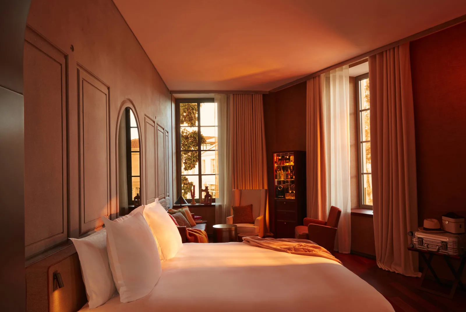 Luxurious bedroom at Mondrian Bordeaux Les Carmes featuring a white bed, warm ochre and red tones, city views through large windows, silk curtains, and contemporary furniture.