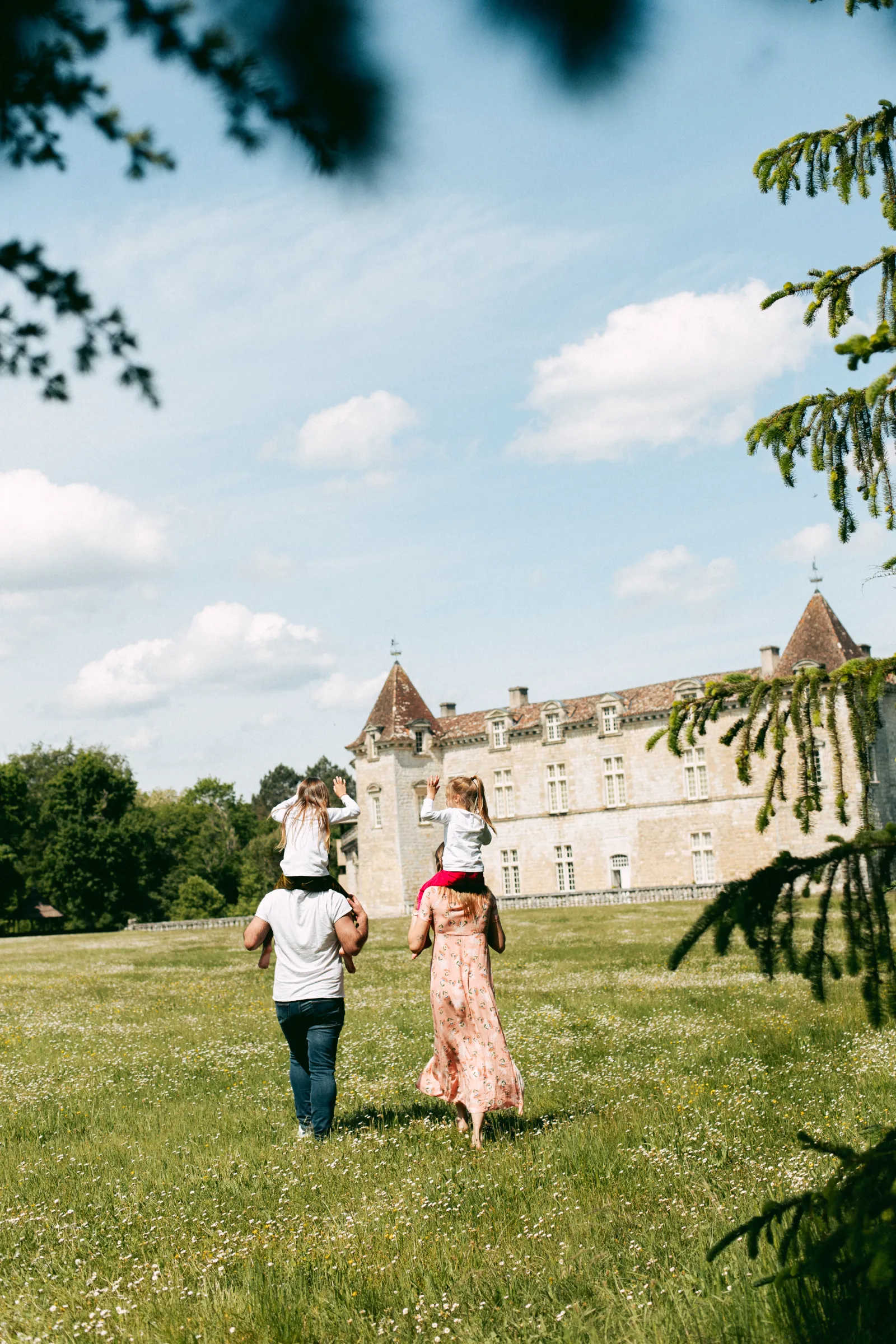 Two adults carrying children on their shoulders walk across a green lawn towards the Royal Castle of Cazeneuve, under a blue sky with white clouds.