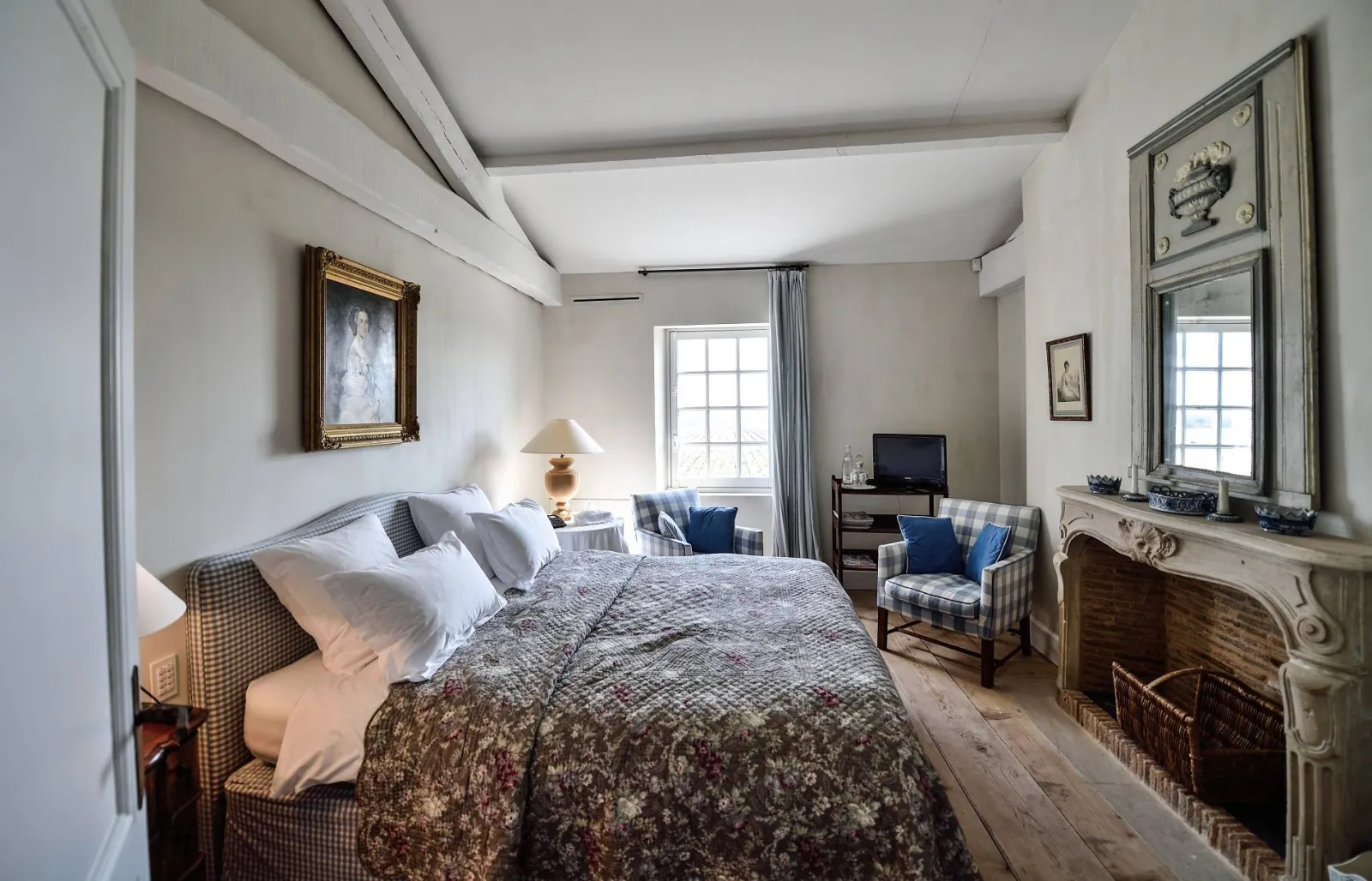 Elegant guest bedroom at Château du Tertre in Bordeaux, featuring a queen-size bed with white linens and patterned bedspread, decorative fireplace, gilt mirror, blue checkered armchairs, and a window overlooking the countryside