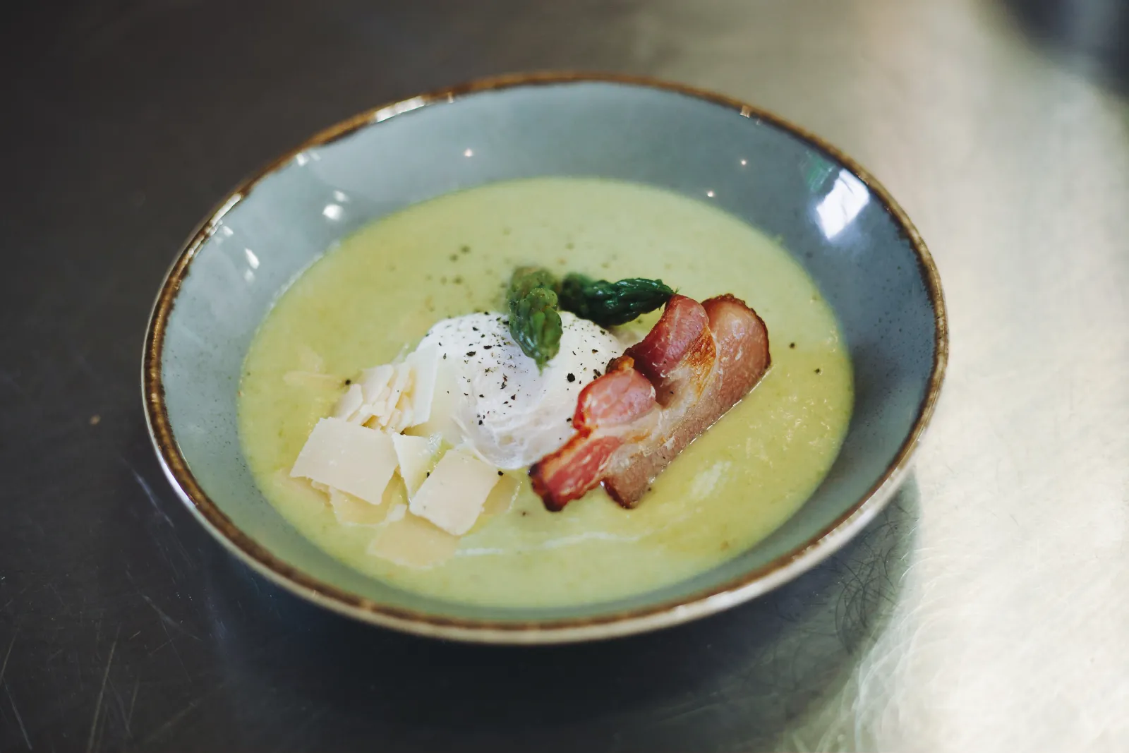 Pale green creamy soup served in a blue-grey bowl, topped with crispy bacon strips, white shredded cheese, a dollop of sour cream, and fresh herbs, presented at Château Ambe Tour Pourret in Bordeaux.