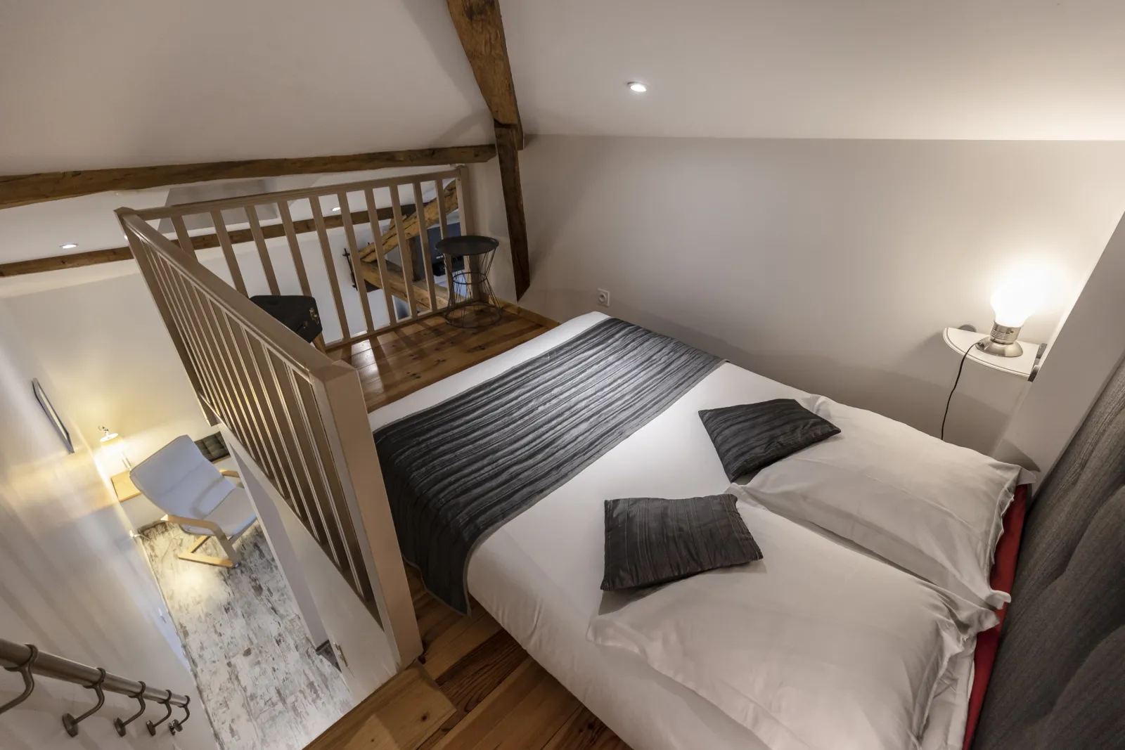 View of a bedroom with sloped ceilings in Studio Merlot featuring a white double bed, black pillows, exposed wooden beams, wooden stairs leading to a mezzanine, minimalist furniture and modern lighting.