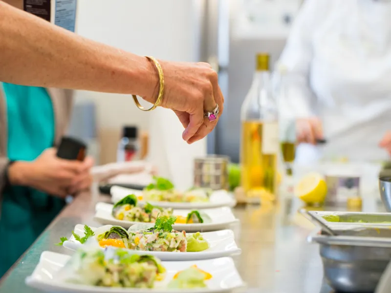 Cooking workshop at Château Kirwan in Bordeaux: a woman in turquoise clothing presents a plated dish garnished with herbs and lemon, with other plated dishes lined up and olive oil bottles in the background.