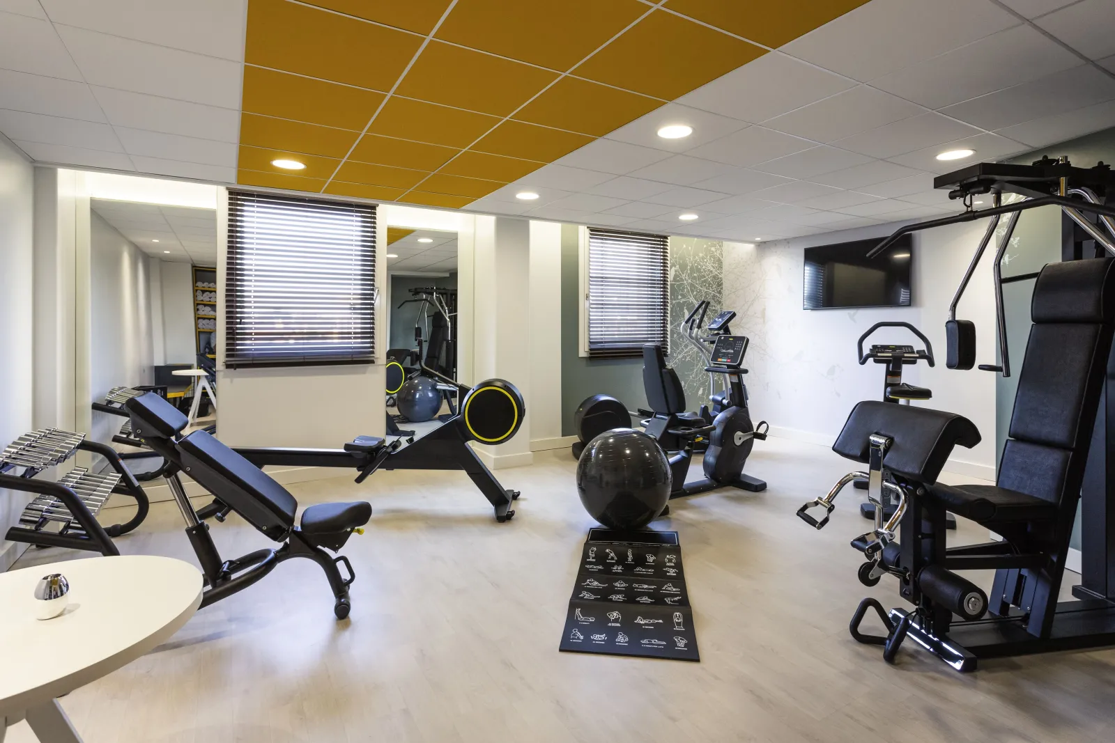 Modern fitness room at Novotel Bordeaux Centre featuring exercise bikes, elliptical machines, treadmills, fitness balls and training equipment, with yellow and white ceiling, mirrors and windows with blinds.