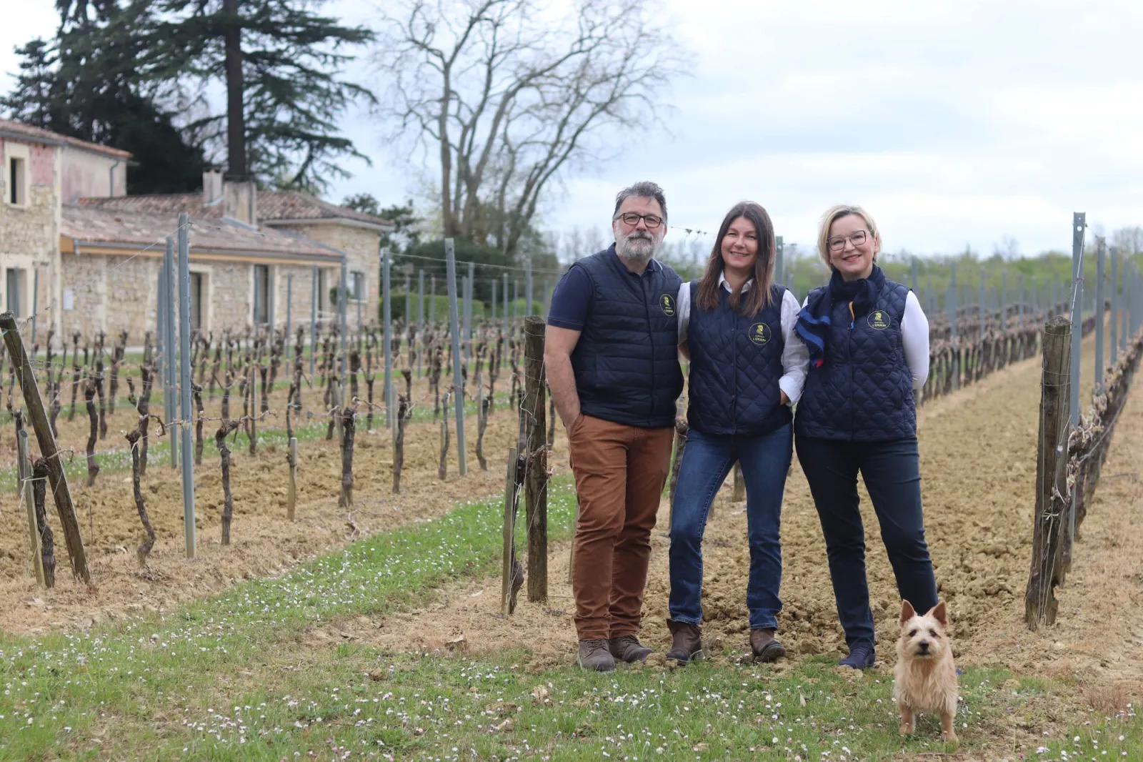 Three people wearing navy vests stand in the vineyards of Château l'Éperon near Bordeaux, with a small white dog beside them and a stone building visible in the background.