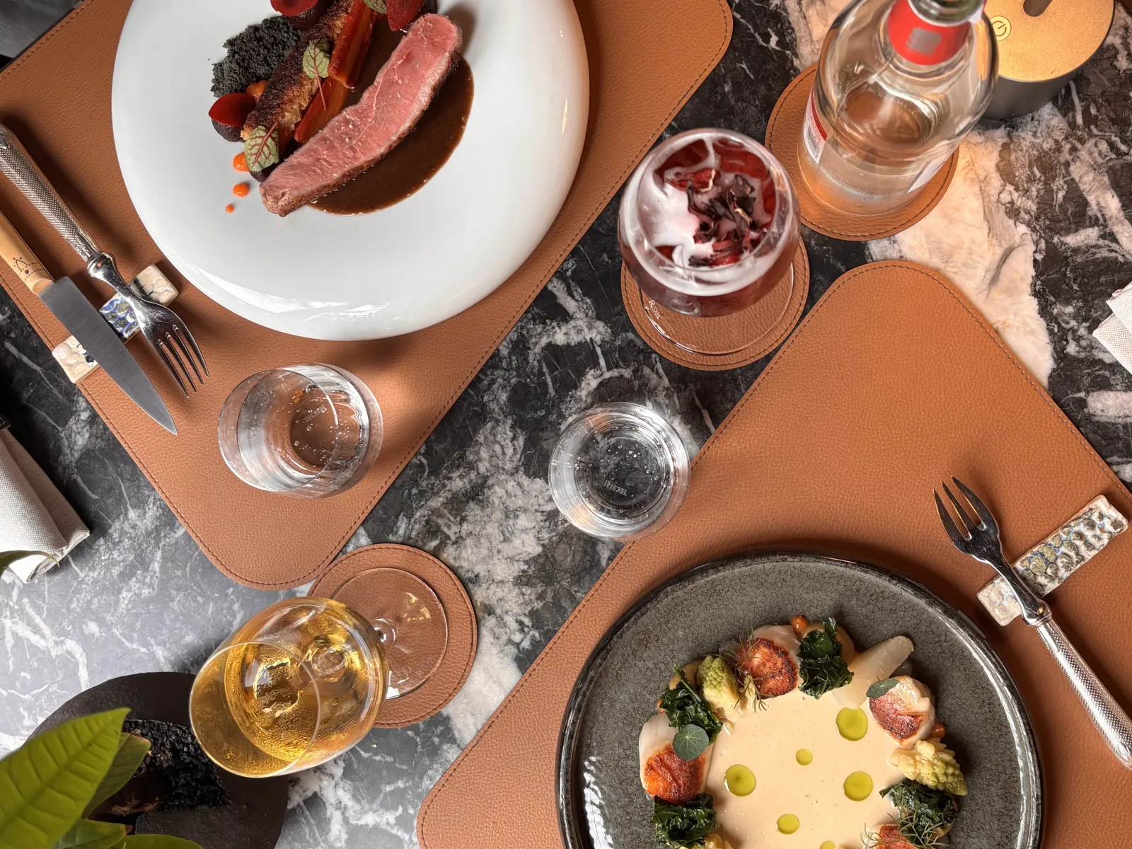 Fine dining table setting with two elegant place settings: white plate with pink meat and delicate garnish, gray bowl with creamy sauce and vegetable decoration, water and wine glasses, cognac leather placemats on gray marble.