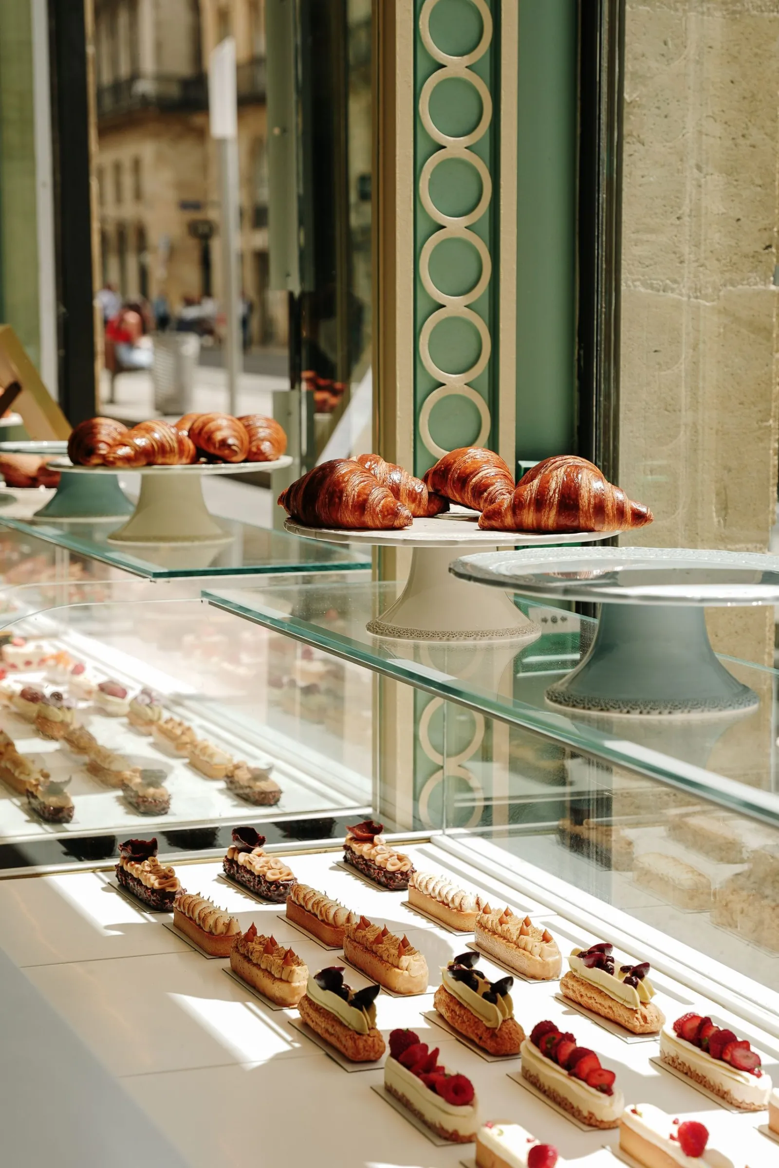 Pastry shop display case featuring golden croissants on ceramic stands and red fruit-topped eclairs in a Bordeaux bakery