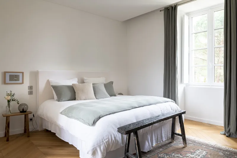Elegant and bright bedroom featuring a king-size bed with white and grey linens, matching pillows, large windows overlooking a garden, wooden nightstand, bench at the foot of the bed, herringbone parquet flooring and grey curtains at Château Toulouse Laut