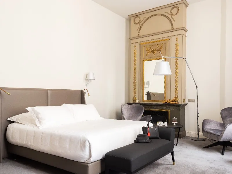 Elegant bedroom at Hôtel Villas Foch in Bordeaux featuring a king-size bed with white linens, a gilded mirror with classic mantelpiece, modern gray armchairs, a floor lamp, and minimalist décor.