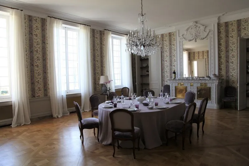 Elegant dining room at Château Gruaud Larose in Bordeaux featuring a round table set for dinner, crystal chandelier, decorative fireplace, floral patterned curtains and Louis XVI style chairs