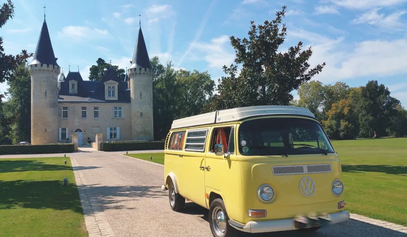 A vintage yellow Volkswagen camper van parked in front of a castle with two pointed towers and turrets, surrounded by green grounds.