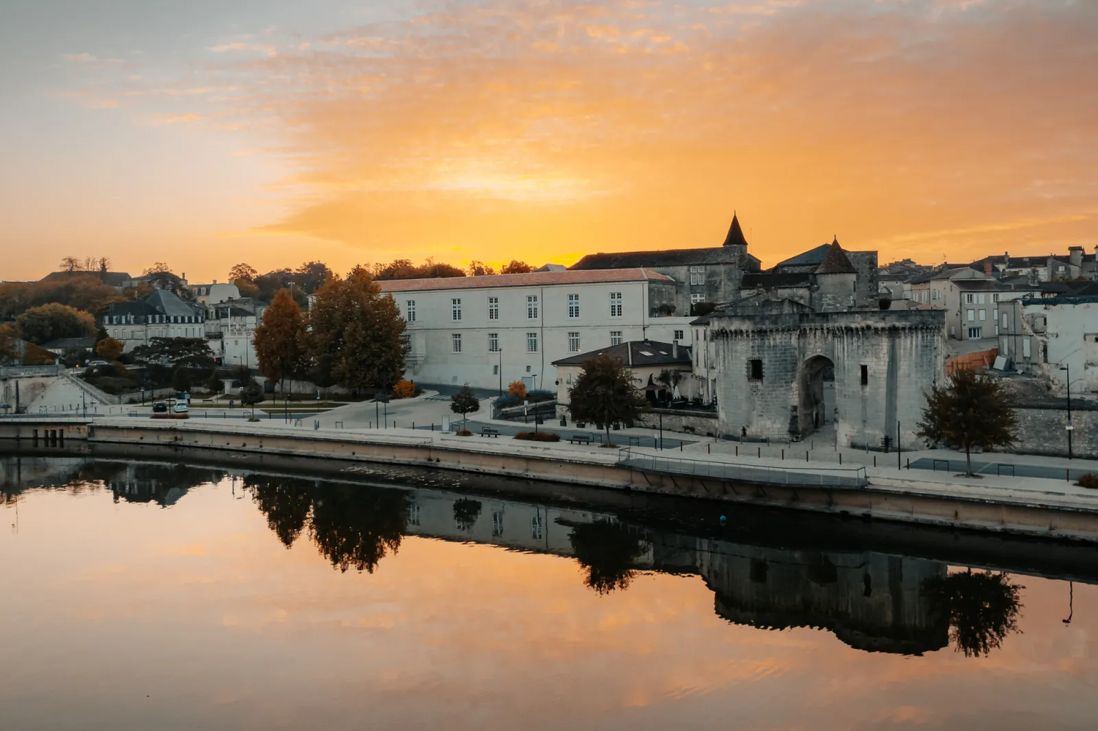 Sunset view of Cognac Castle featuring medieval towers and historic buildings reflected in the Charente River, surrounded by fortified walls and ancient architecture.