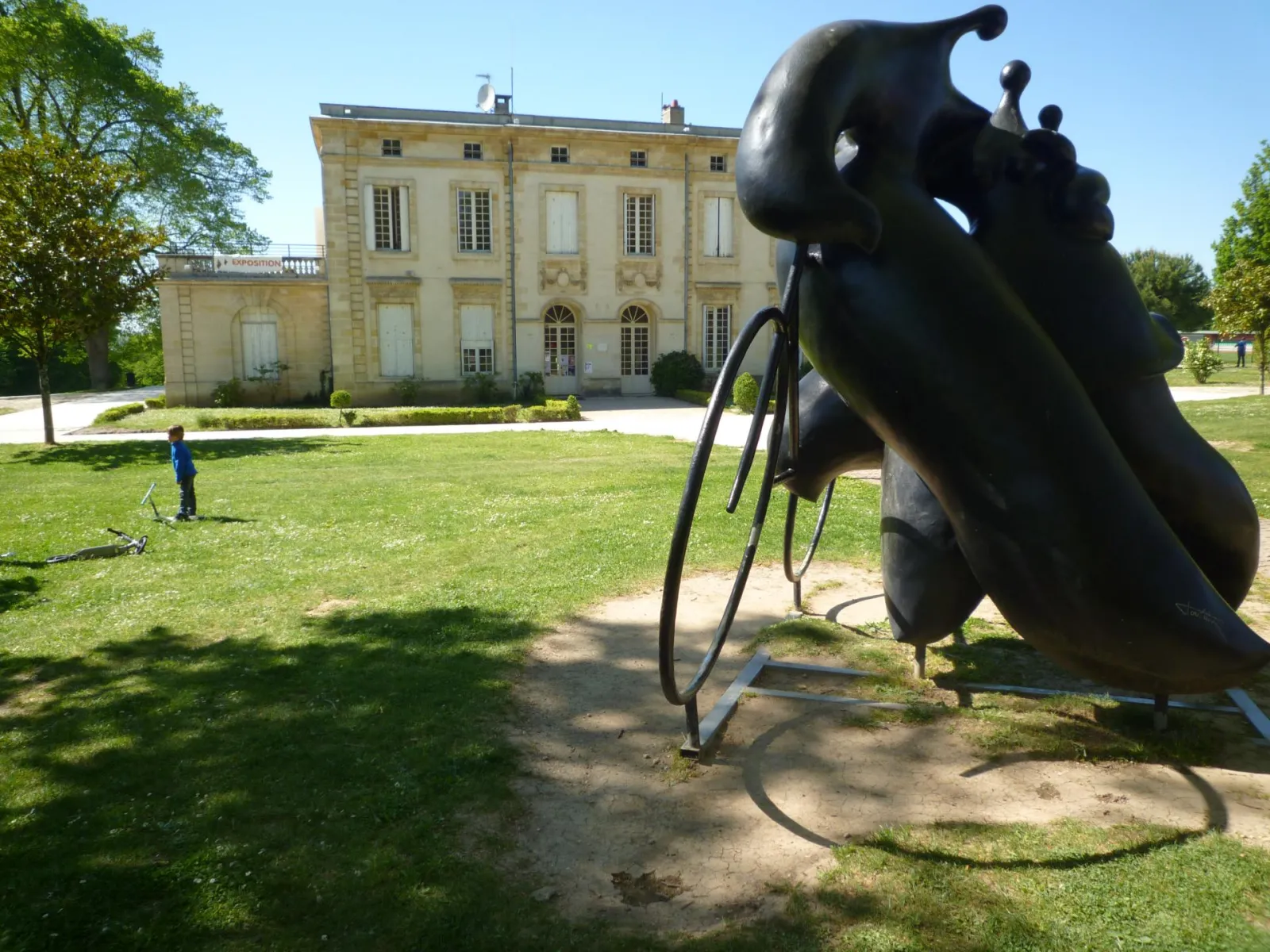 Black abstract sculpture in the foreground with the neoclassical Palmer mansion in the background, a beige building with regular windows set in a green park in Bordeaux, with a child playing on the lawn.