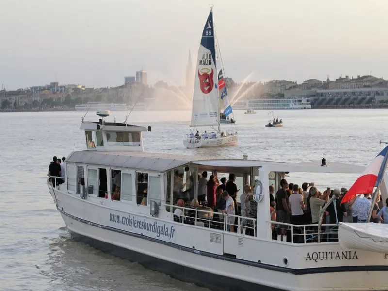 Aquitania cruise boat from Croisières Burdigala sailing on the Garonne river in Bordeaux with passengers aboard, a sailing boat in the background, and the city waterfront visible.